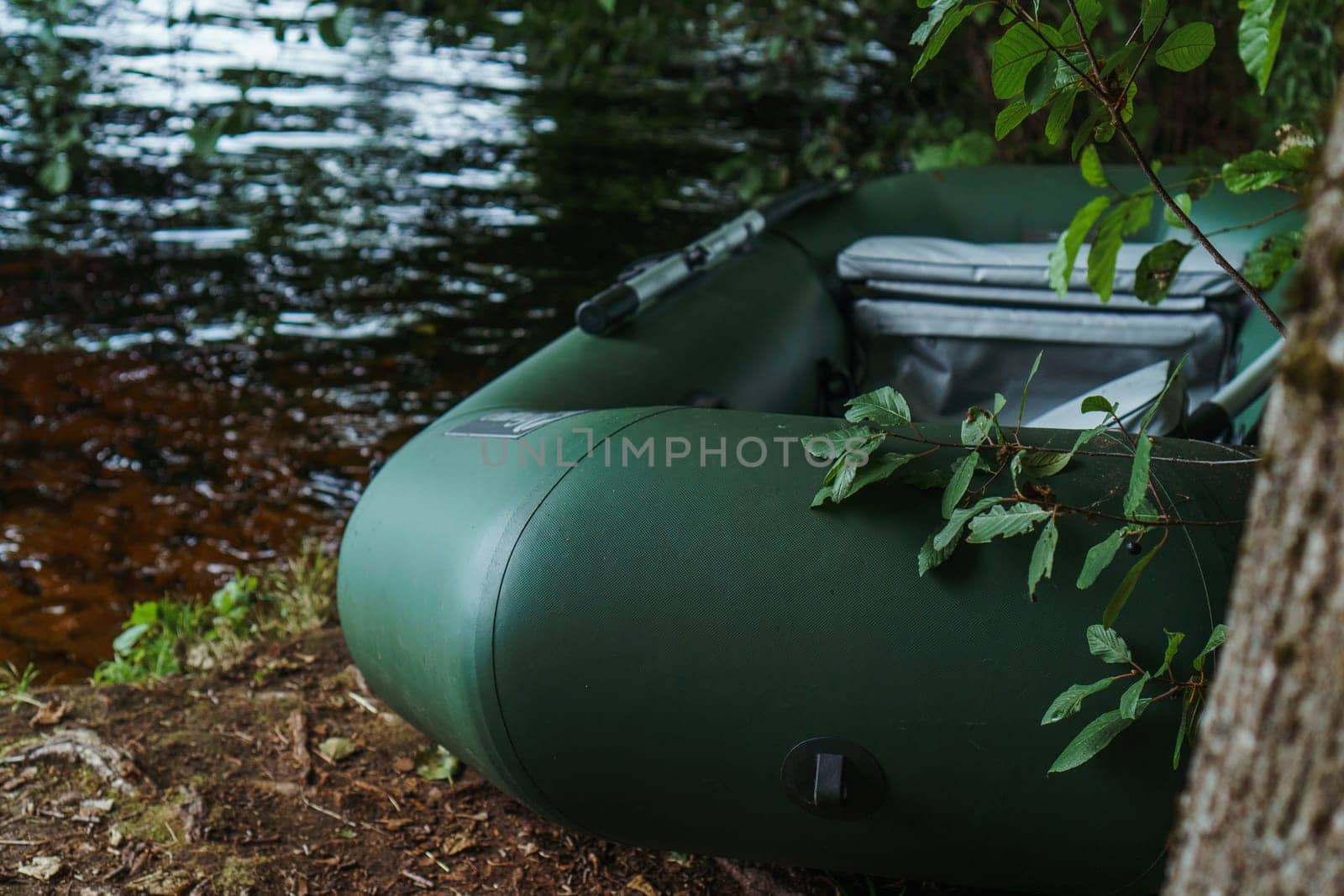 Modern green inflatable boat parked on shore near lake in evening, standing close to tree, slightly covered by branches and leaves. Transportation for floating and relaxing on water in rural site.