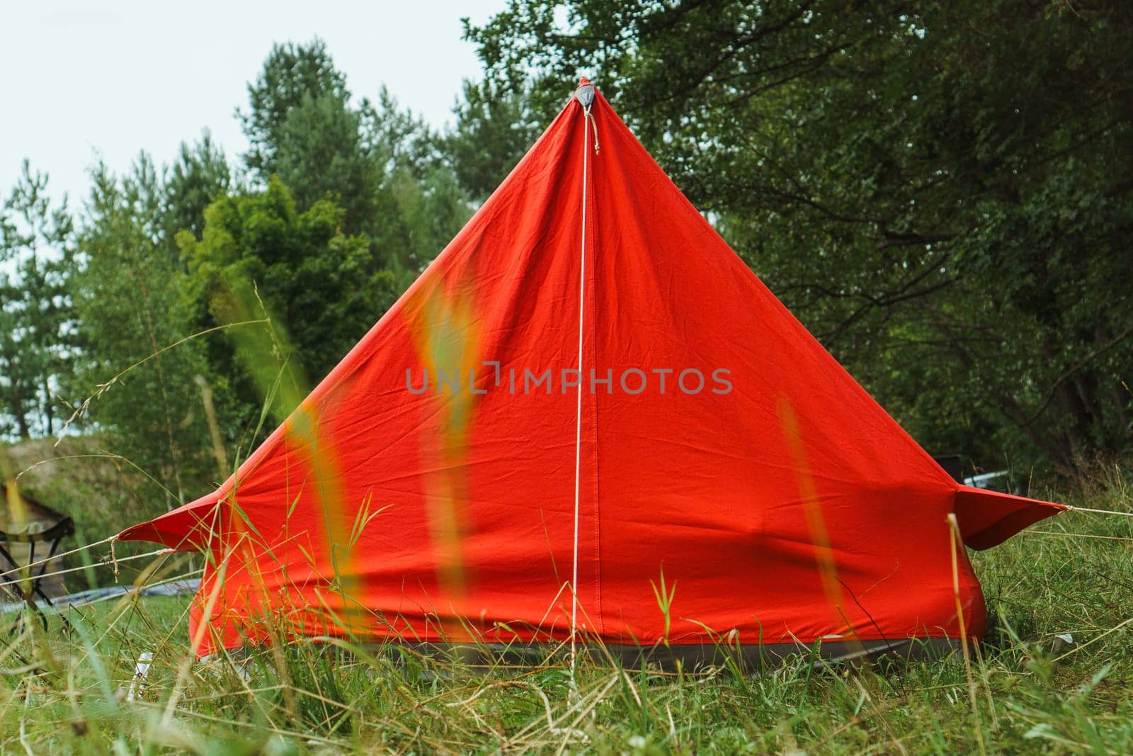 Big red camping tent constructed for relaxation on green lawn near forest in non-urban place. Charming bright shelter pitched on grass in summer day, attached with ropes and ready for night.