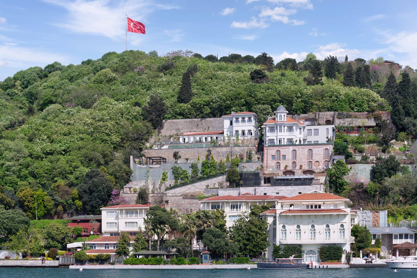 Group of traditional houses on a green hill by Bosphorus in Istanbul, Turkey, with a large Turkish flag flying overhead. The trees in the foreground are lush and green. The sky is clear and blue