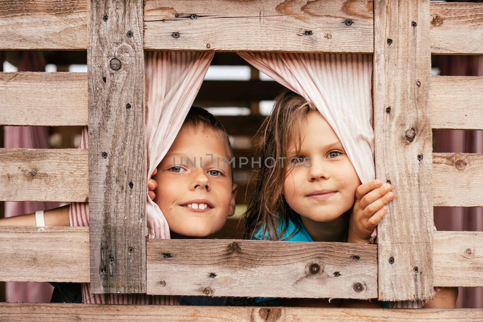 Two charming children, boy and girl, look out small window of wooden children's house. Holidays in village. Happy childhood. Children's games and entertainment. Children's emotions.