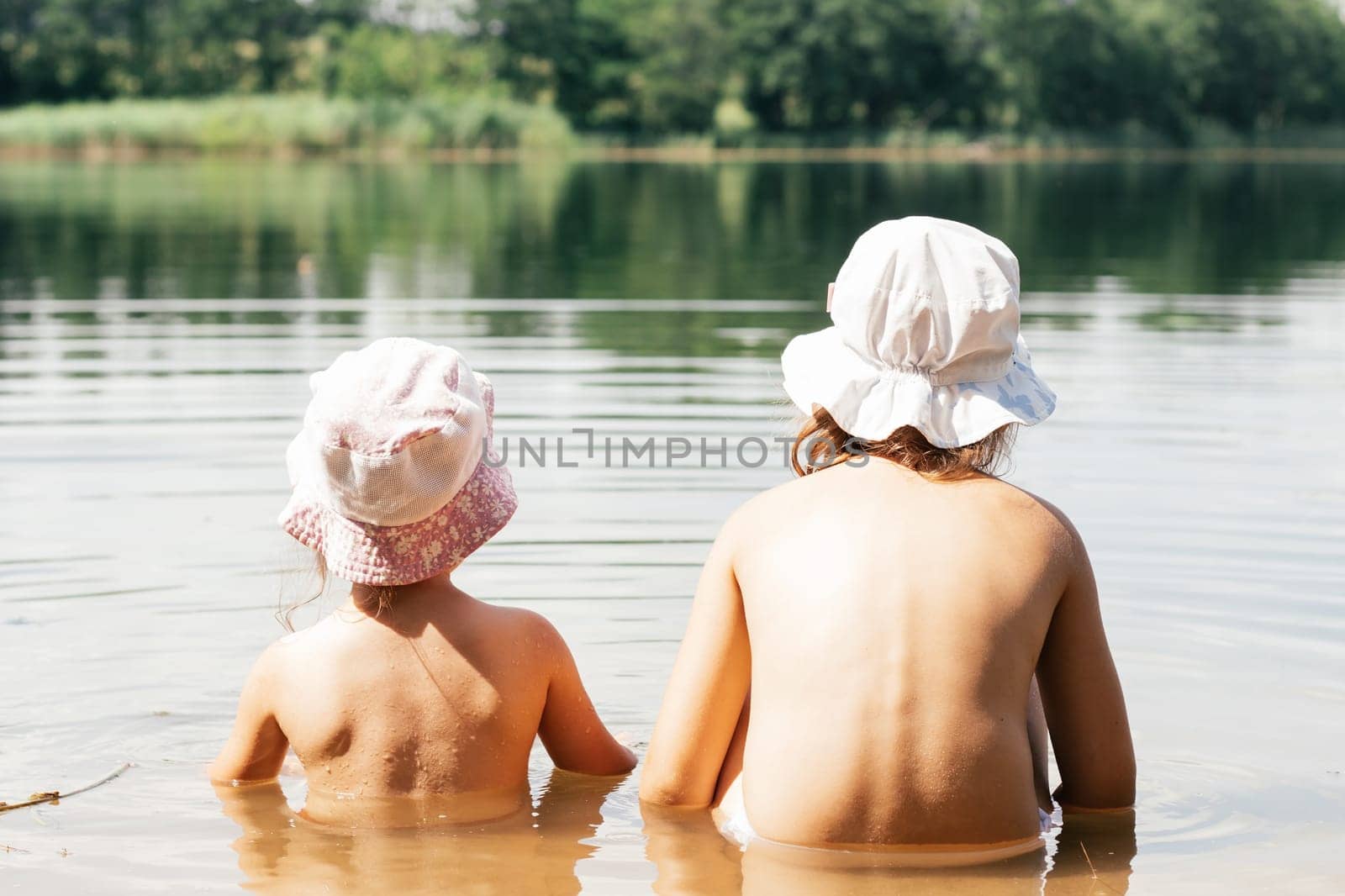 Two children, brother and sister, friends in summer panama hats are sitting in lake in water with their backs to camera. Holidays in village. Happy childhood. Children's games and entertainment.