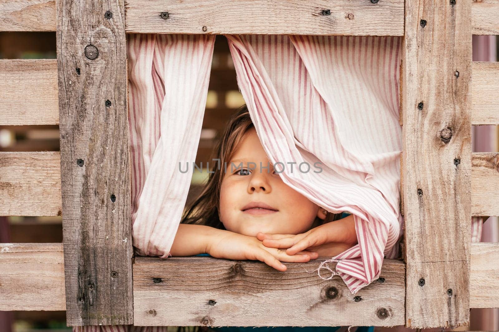 Happy little girl looks out window of wooden children's house. Holidays in village. Happy childhood. Children's games and entertainment. Children's emotions. Positive. Imagination and fantasy.