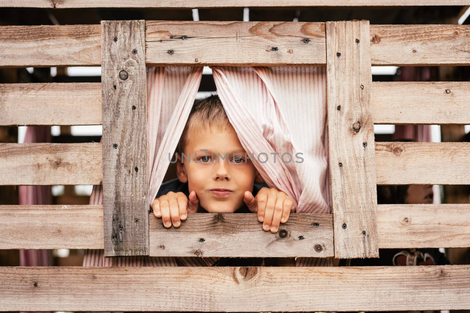 Cute little boy looks out window of wooden children's house. Holidays in village. Happy childhood. Children's games and entertainment. Children's emotions. Positive. Imagination and fantasy.