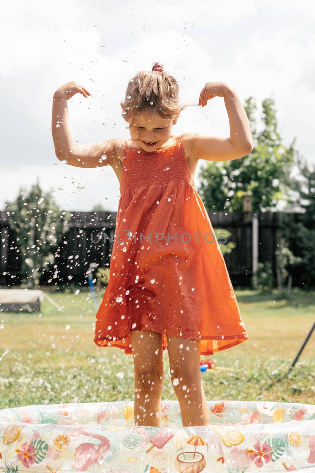 Charming little girl in orange sundress is standing in inflatable pool in spray of water, laughing on summer day outside. Games and entertainment. Summer holidays. Happy childhood.Happy. Positive.