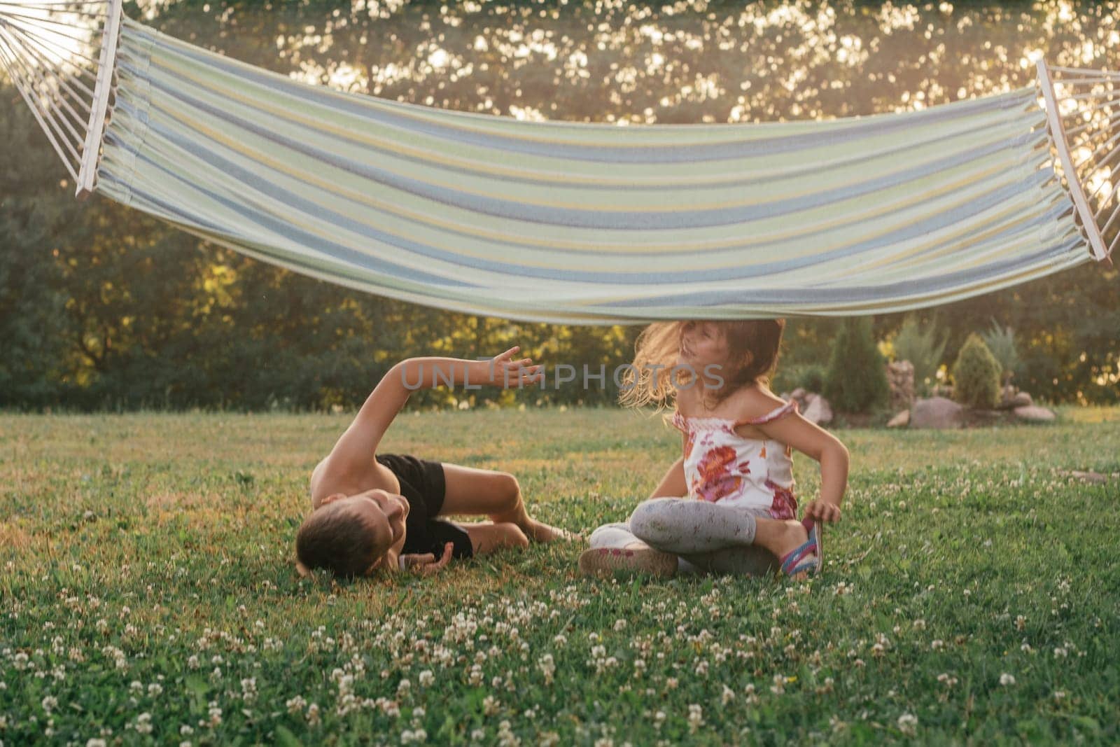 Two children, boy and girl, brother and sister play on grass under hanging hammock on sunny summer day in nature. Summer holidays in village. Games and entertainment.Happy childhood. Positive .