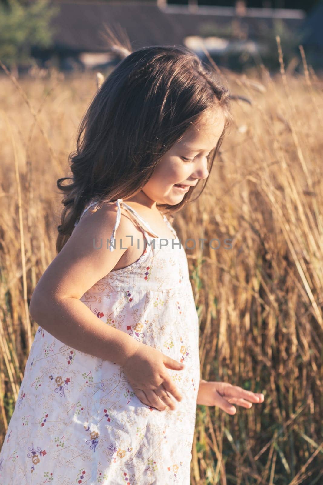 Close-up. Portrait of happy smiling little girl in light sundress made of natural fabric on summer day in field among tall cereals. Summer holidays in village. Children's emotions. Happy childhood.