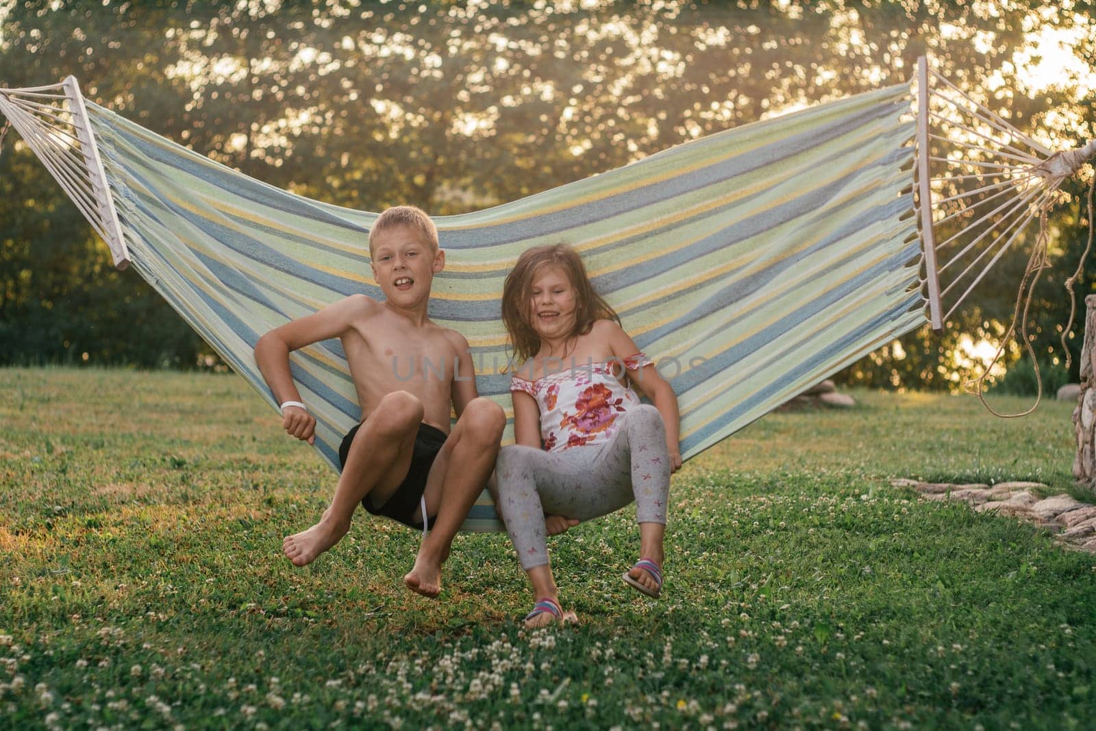 Happy children, boy and girl, brother and sister, friends laugh, swing in hammock on summer day in nature. Summer holidays in village. Games, entertainment. Positive emotions. Childhood friendship.
