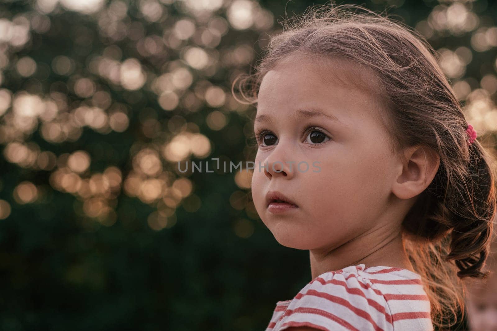 Close-up. Portrait of cute serious little girl looking to side against background of unfocused trees at sunset in summer. World through eyes of child. Children's emotions. Childhood.