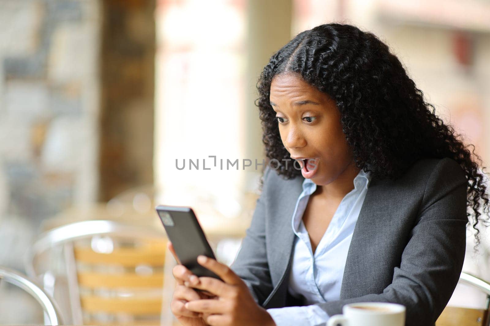 Amazed black executive checking smart phone in a bar terrace