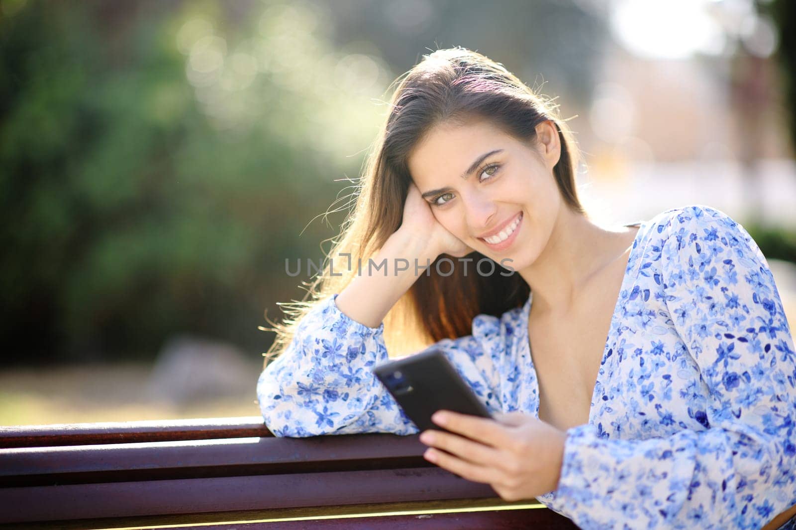Happy woman looks at you holding phone sitting in a park