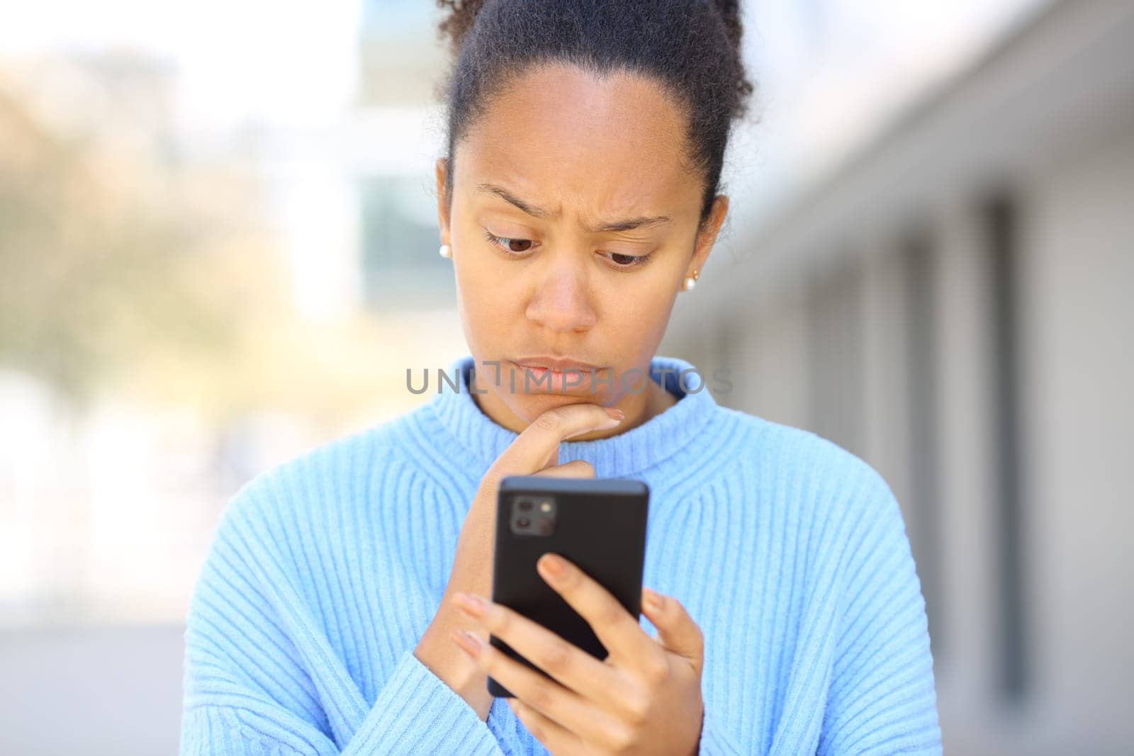 Front view portrait of a worried black woman checking phone in the street