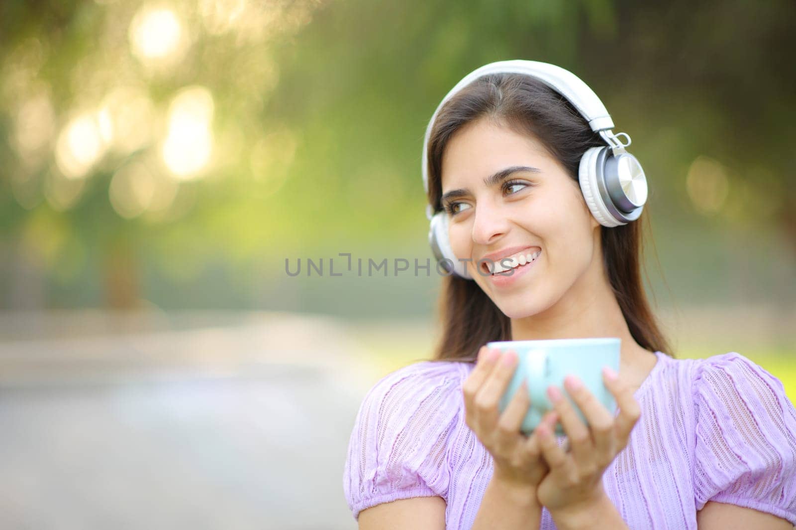 Happy woman wearing headphone laughing and listening music and drinking coffee in a park