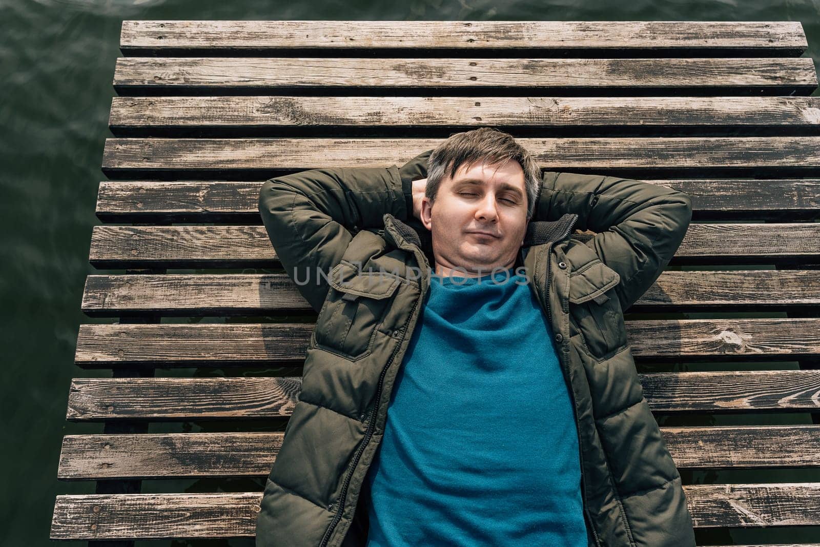 Top view of a man lying on a wooden boardwalk boat dock on a lake with his eyes closed in a jumper and jacket.