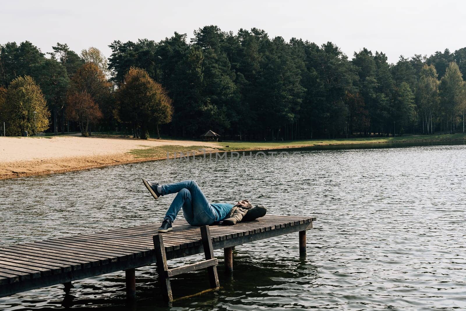 A man rests lying on a wooden pier on a lake in jeans and a jacket on an autumn day.