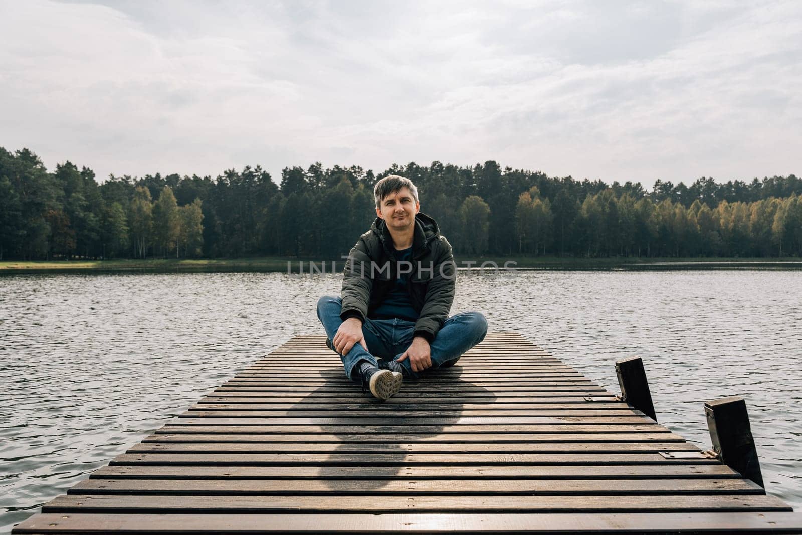 A man enjoys a sunny autumn day while sitting on a wooden pier on a lake.