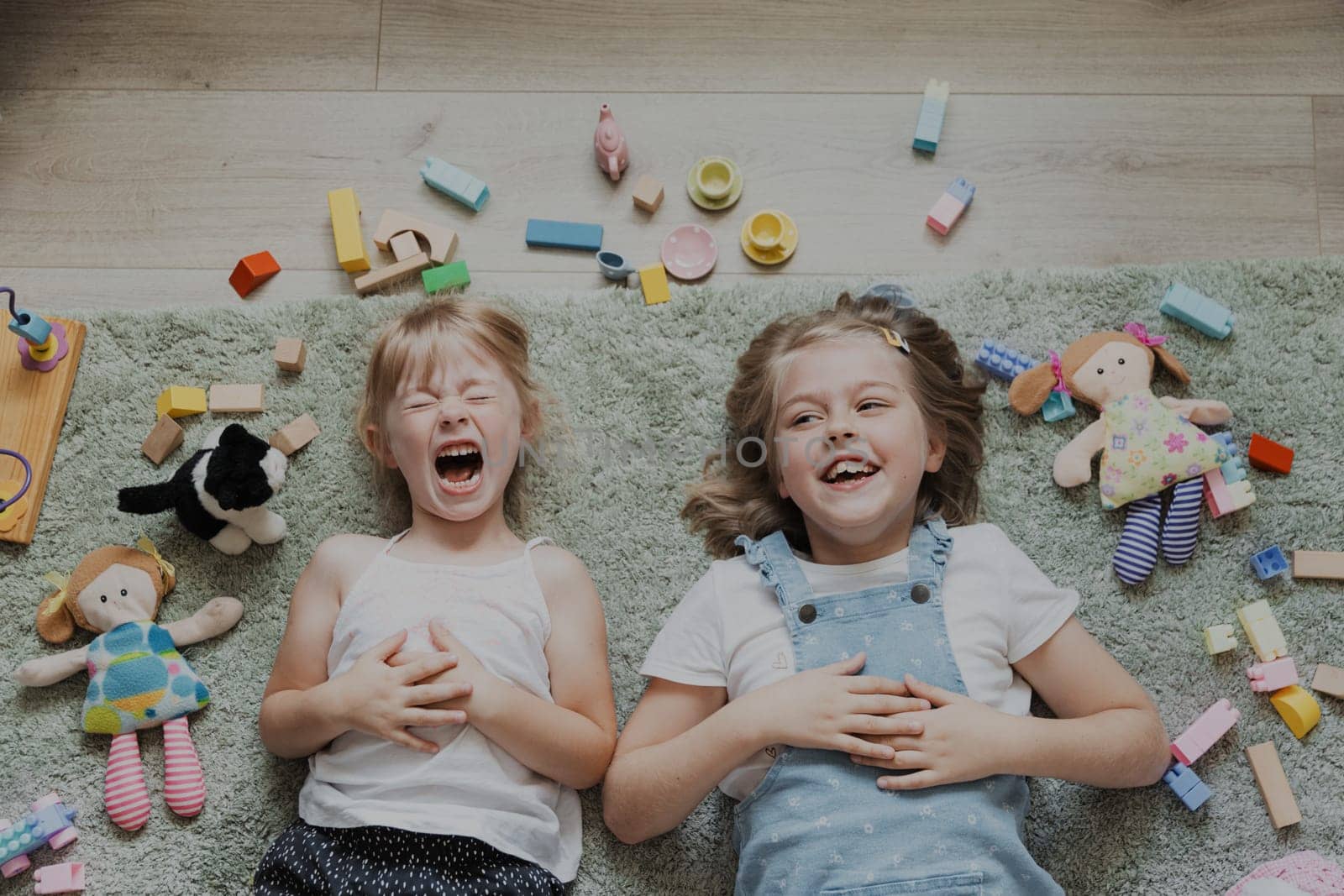 Top view from above on children playing with colorful toys. Portrait of kids lying on carpet at home bedroom or kindergarten. Two happy smiling sisters. Educational eco-friendly games
