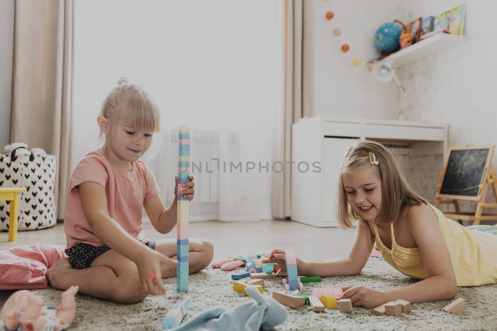 Children playing with colorful toys. Kids sitting on carpet at home bedroom or kindergarten, nursery. Educational eco-friendly game for child. Cute little sisters build house, tower, having fun