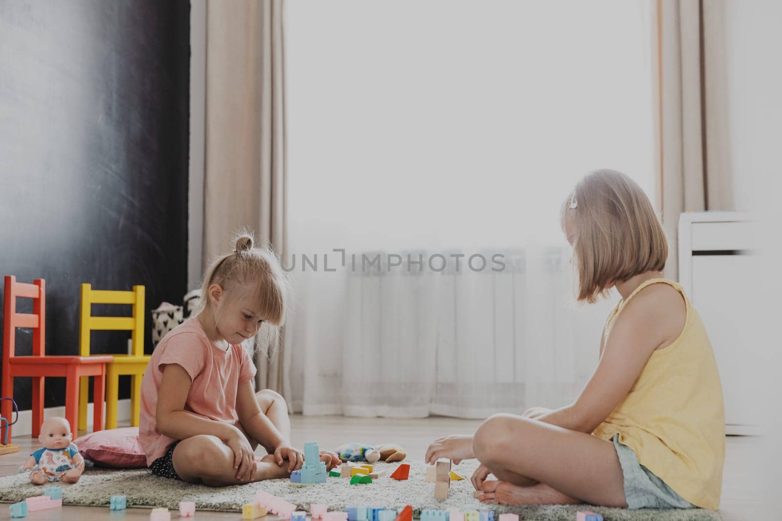 Children playing with colorful wooden toy building blocks, bricks. Kids sitting on floor at sunny white home bedroom or kindergarten, daycare playroom. Funny educational eco-friendly game for child