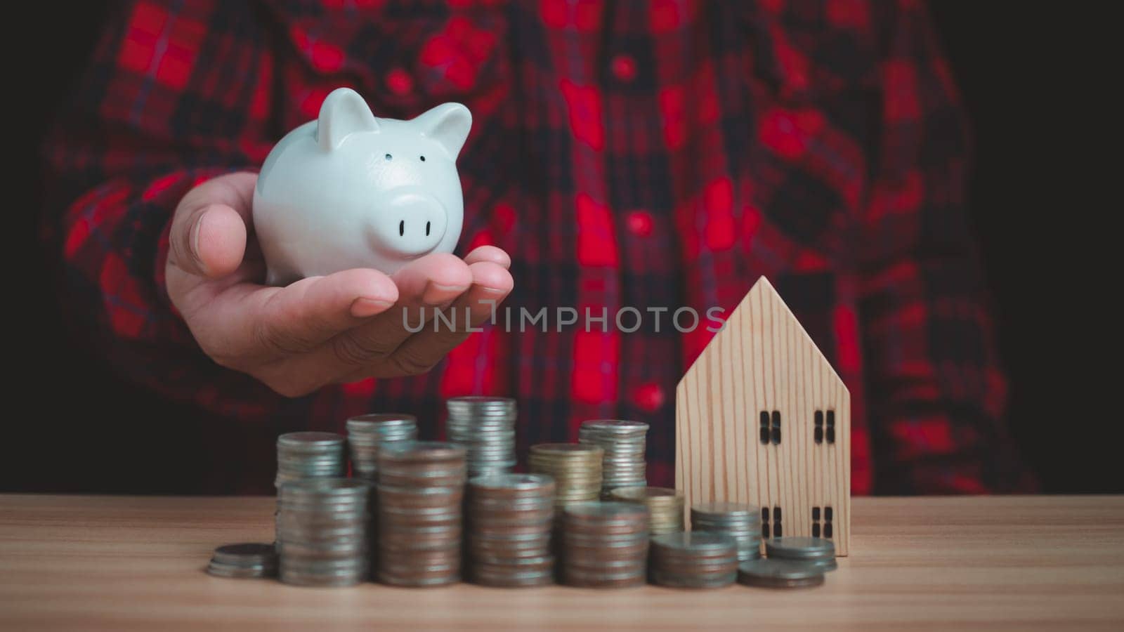 Human holds a piggy bank and there are coins and model wooden houses lined up on the wooden floor. Concepts of finance, savings, investment, and setting goals for fixed deposits with banks.