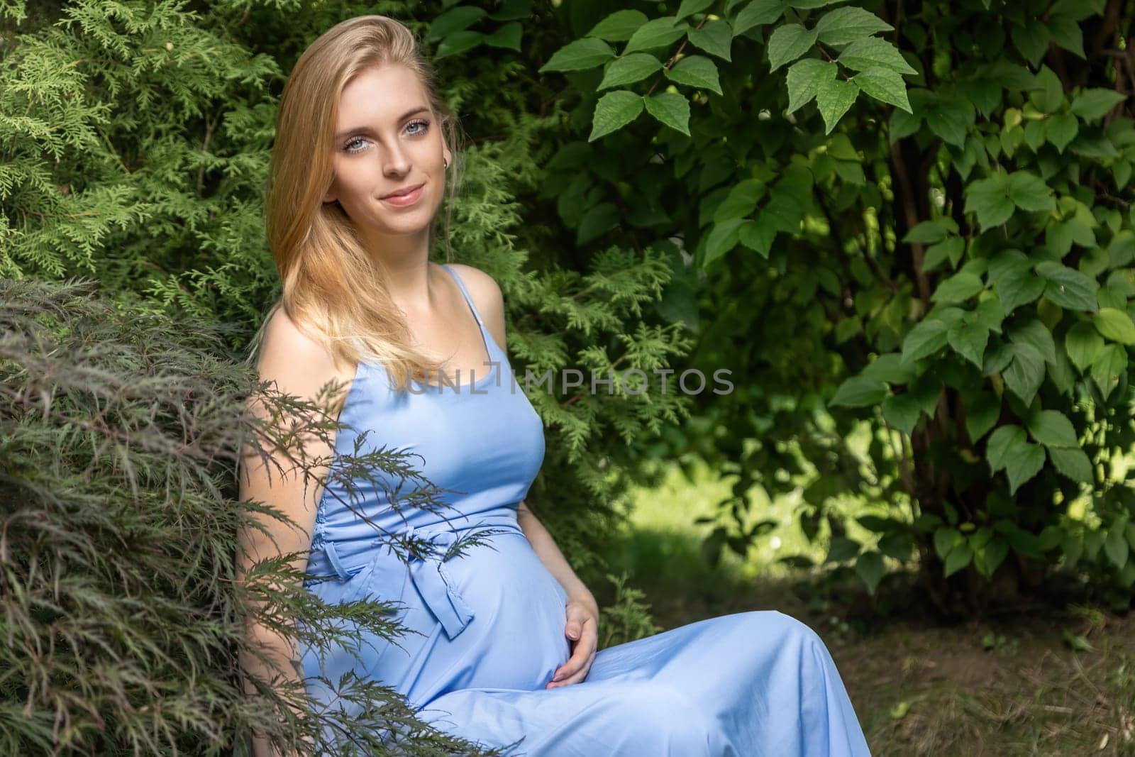 Pregnant young blonde woman is posing outdoors looking at the camera.  Horizontally.