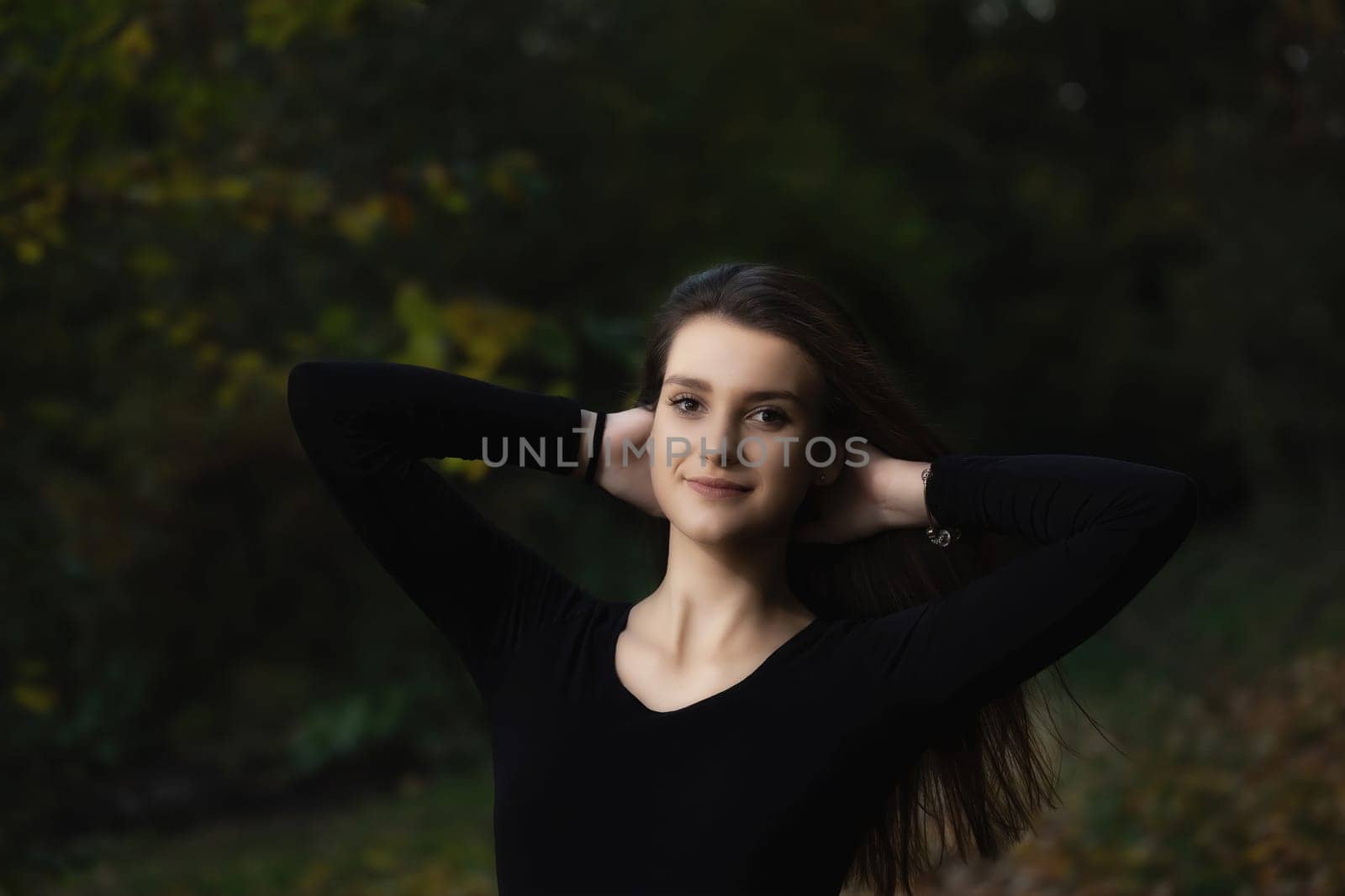 Portrait of attractive long haired young woman posing with hands behind head outdoors on dark background.  Horizontally.