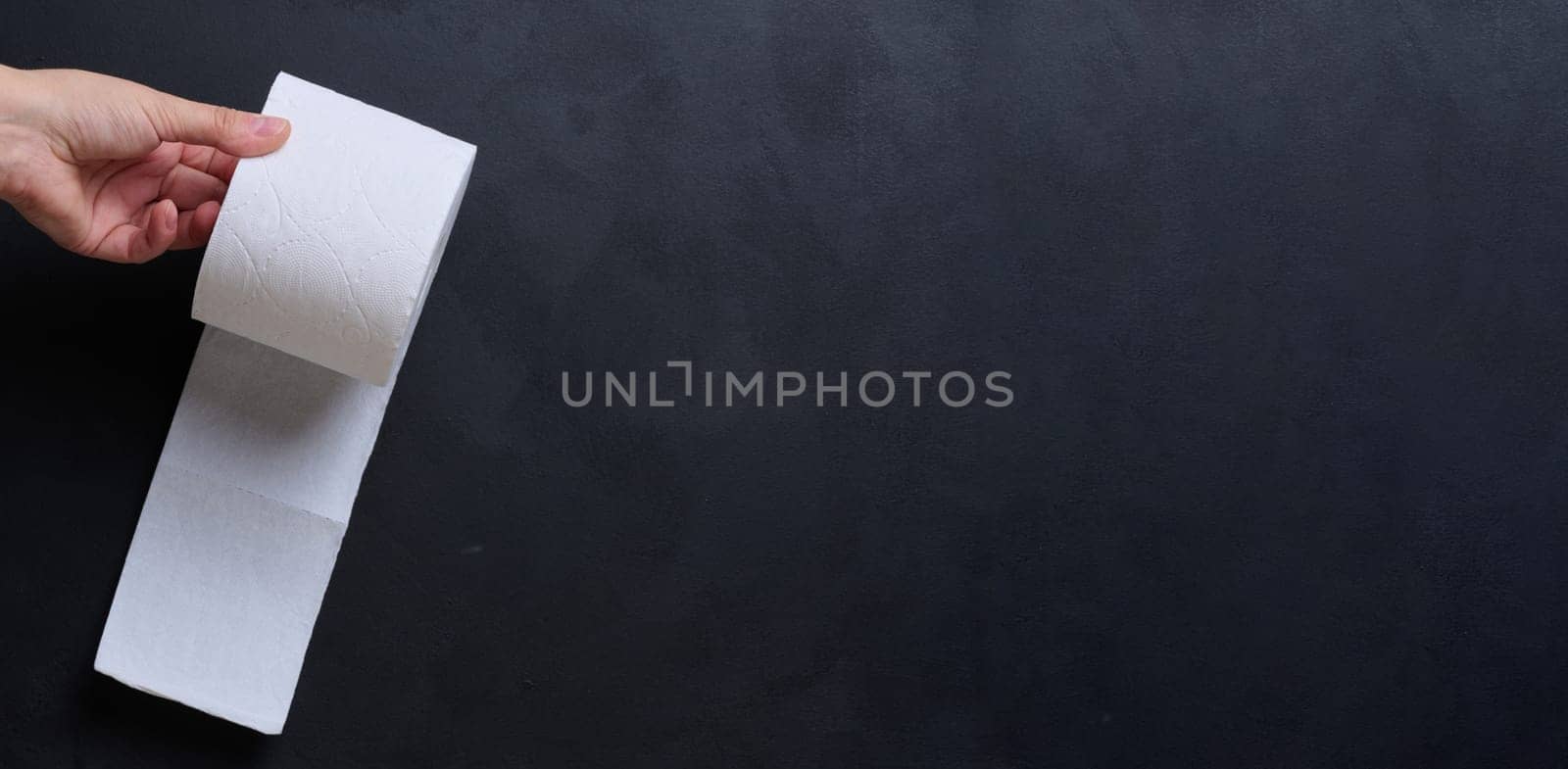 A female hand holds a roll of white toilet paper against a black background. Copy space