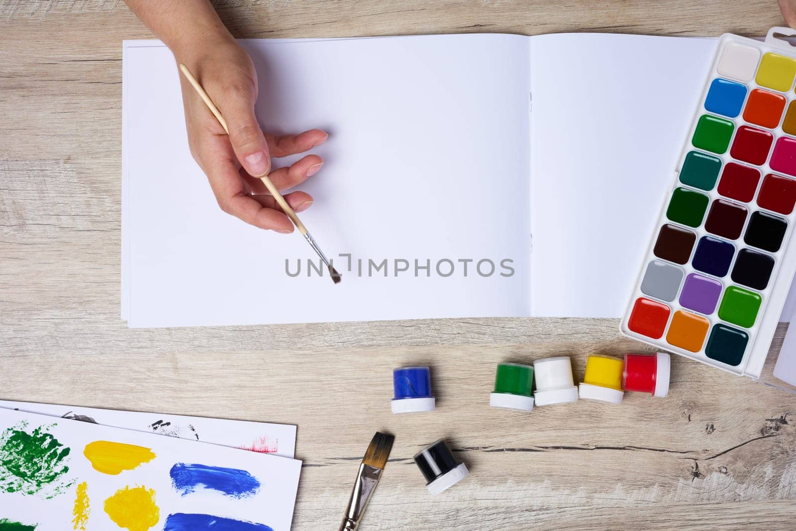 Set of watercolor paints, brushes and paper with different strokes and colors. Female hands holding a brush