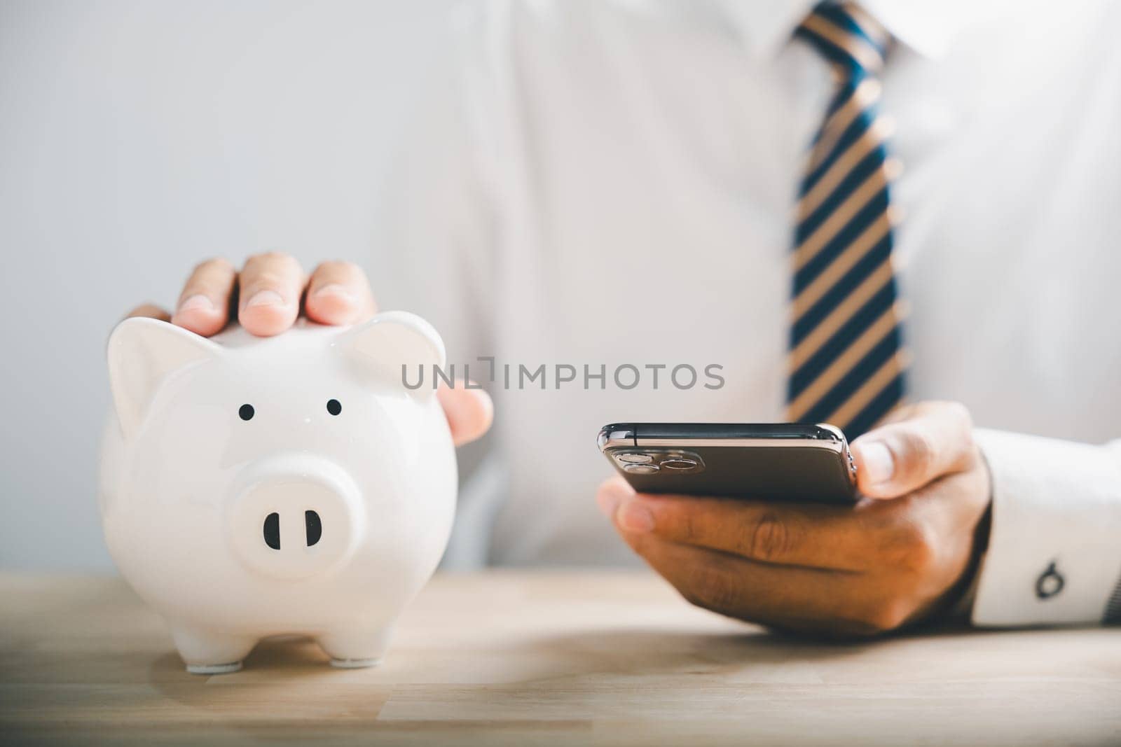 Mobile banking and budgeting, man saves money in piggybank. Growing fund concept on white desk. Financial support banner. Smartphone and piggybank illustrate wise investment. Financial Money Advice