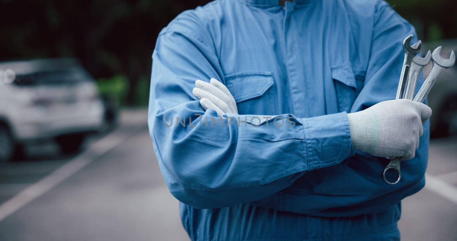 Close up of professional mechanic arm cross while holding wrench and wear blue uniform. Mechanic holding a spanner getting ready to work on a car at his workshop. Car mechanic proudly posing.