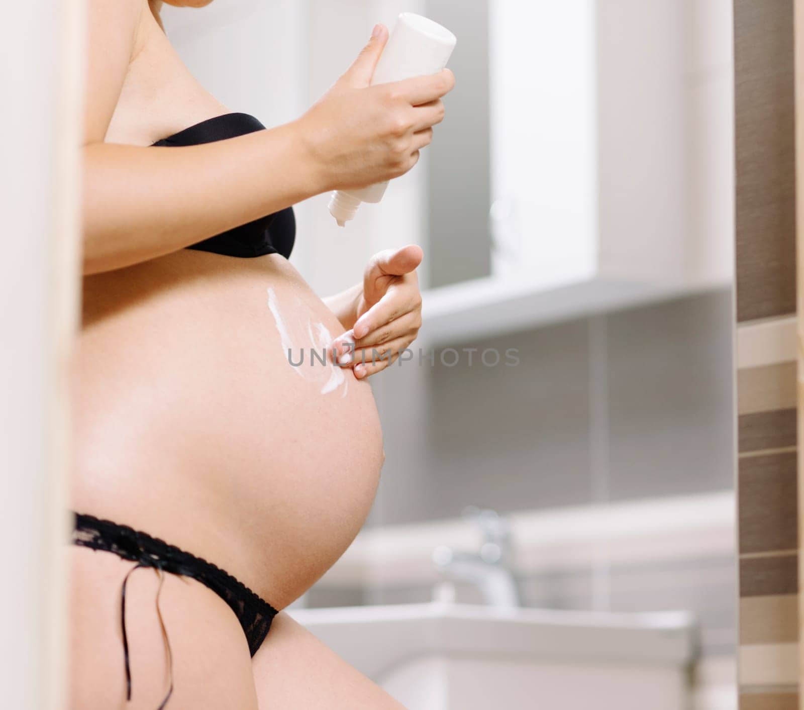 Pregnancy skin care for stretch marks. Beautiful pregnant woman in bathroom applying cream on her belly skin. Maternity cosmetics and skincare products.