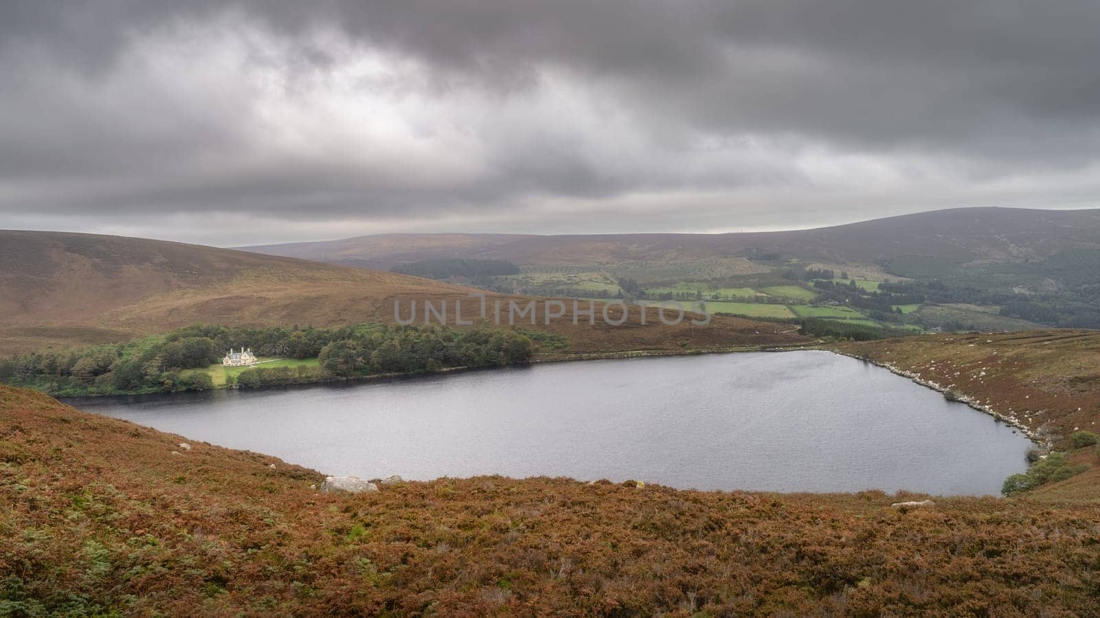 Mansion or house hiding in small forest at the edge of lake Lough Bray, surrounded by dramatic storm clouds. Hiking in Wicklow Mountains, Ireland