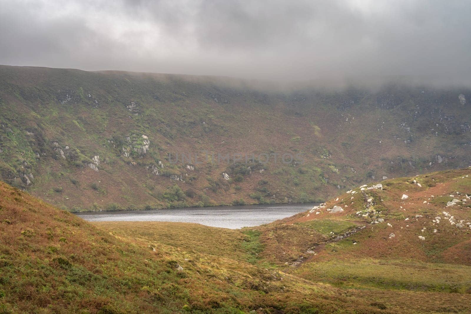 Lake, Lough Bray hiding behind hills and cliffs, surrounded by dramatic storm clouds and sky. Hiking in Wicklow Mountains, Ireland