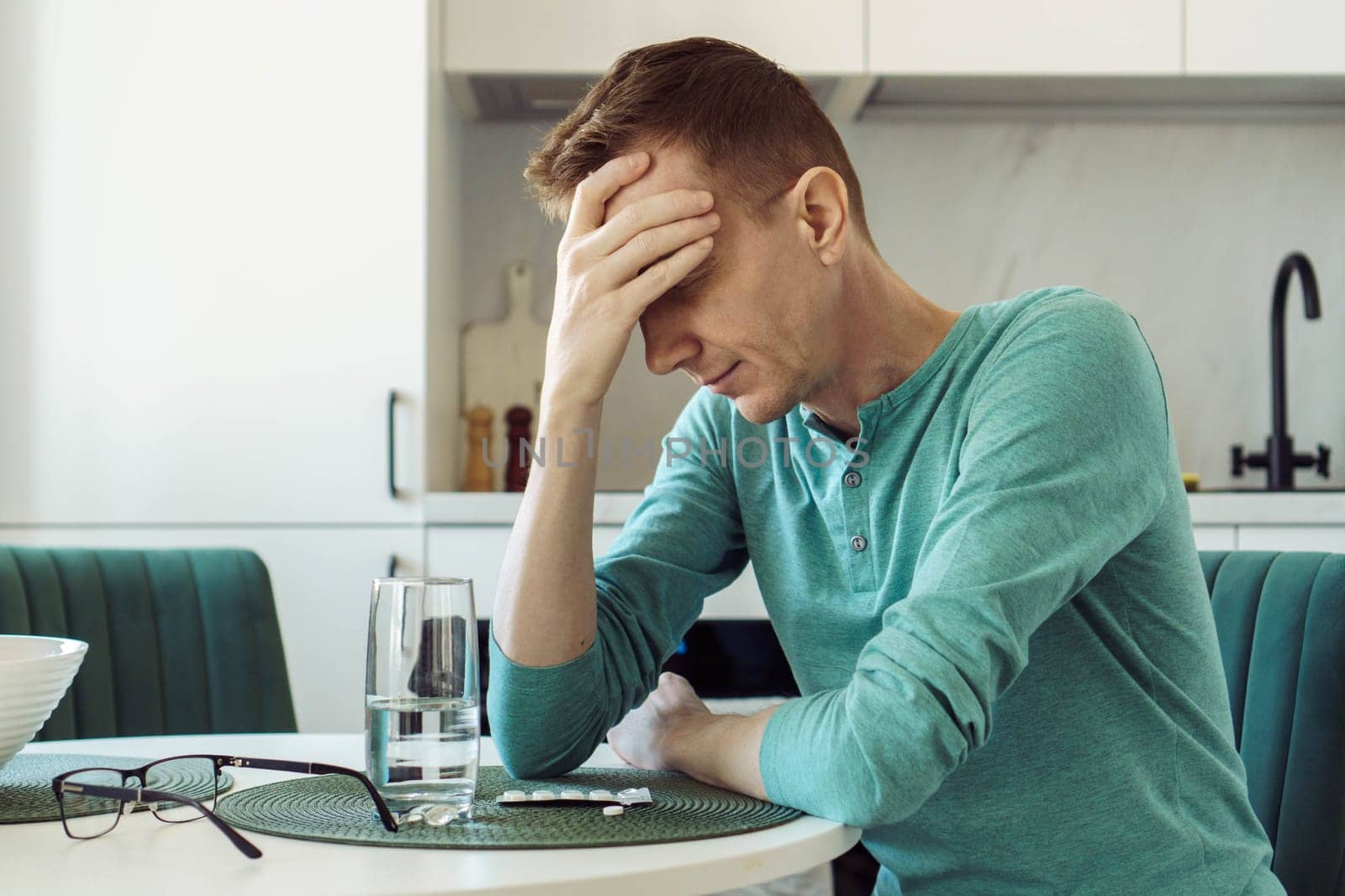 Man in turquoise T-shirt is sitting at home at kitchen table and holding his head with his hand. Illness and loneliness. On table is glass of water and pills, painkillers, antidepressants.