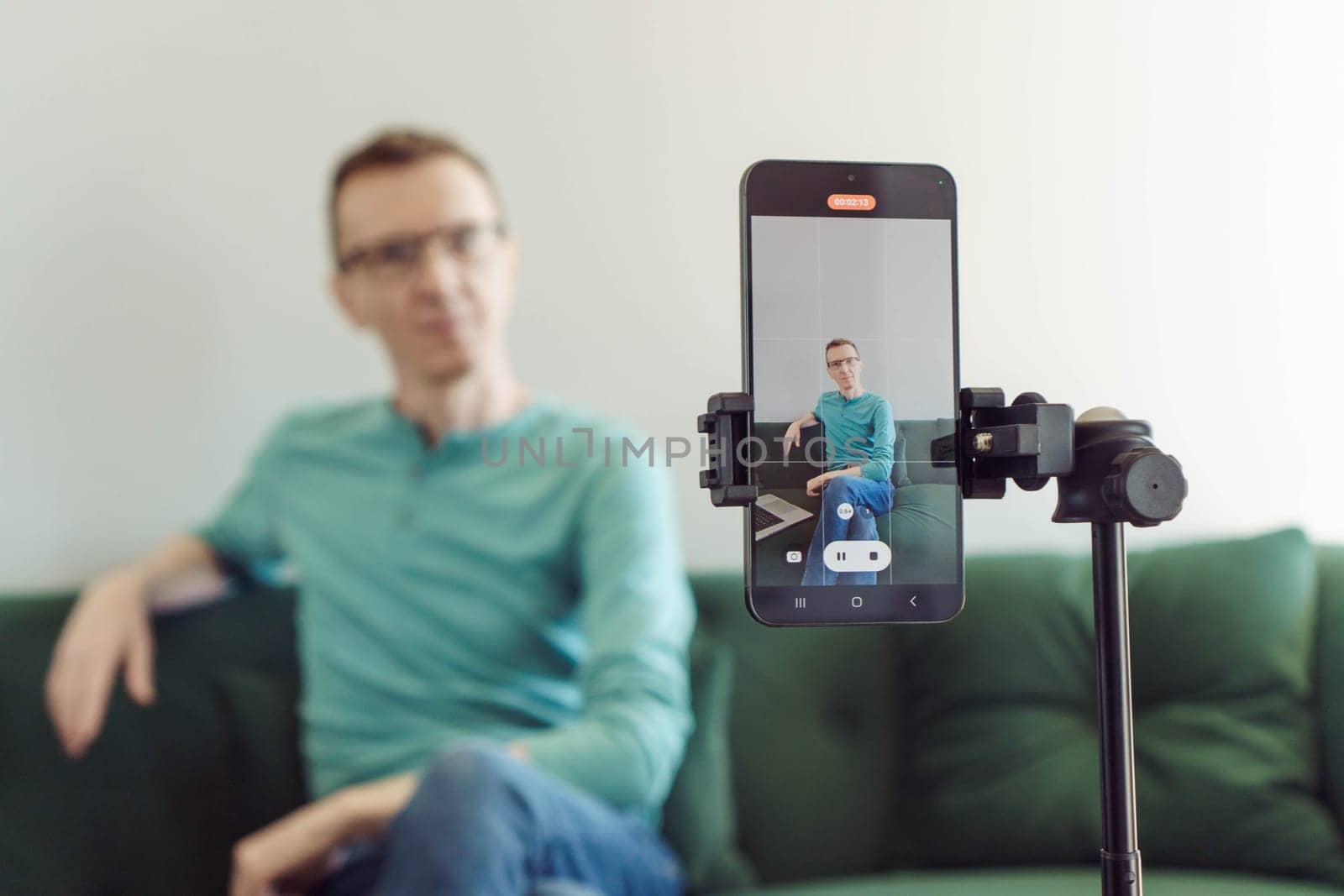 Unfocused man is sitting on sofa in background, focusing on phone on tripod with picture of camera shooting. Blogger. Modern technologies. Online training. Video recording of trainings. Video chat.