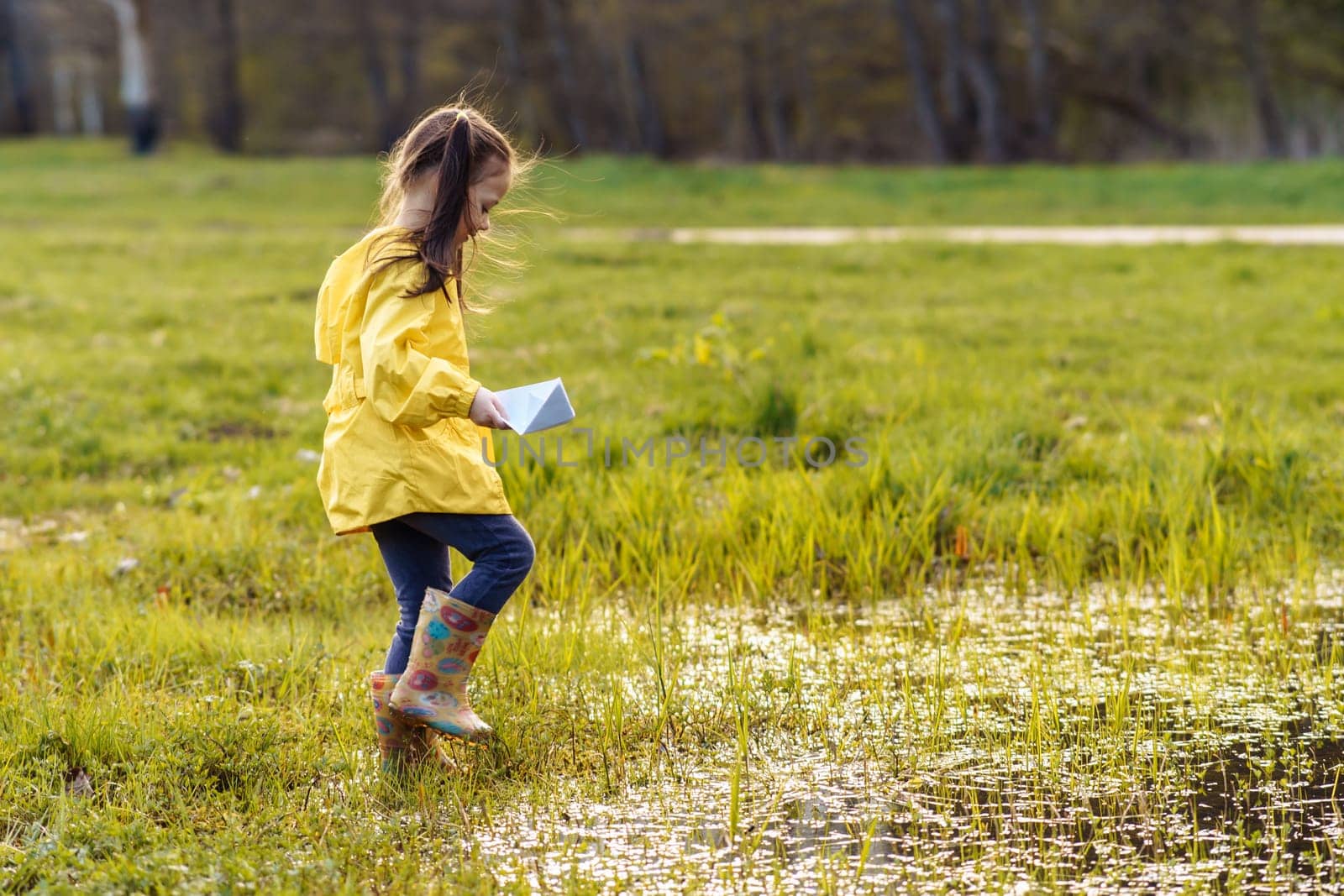 Adorable girl in bright yellow jacket and rubber boots walking in puddle on green lawn, carrying white paper ship in hand and preparing to launch boat on water, spending autumn day in nature.