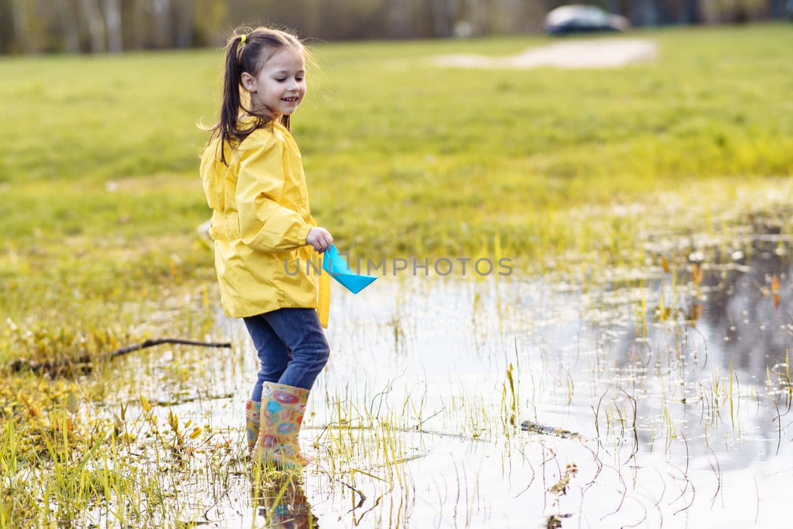 Happy female kid, wearing yellow jacket and rubber boots, standing in puddle on green site, holding blue paper ship in hand and turning head over shoulder with soft smile, enjoying game on fall day.