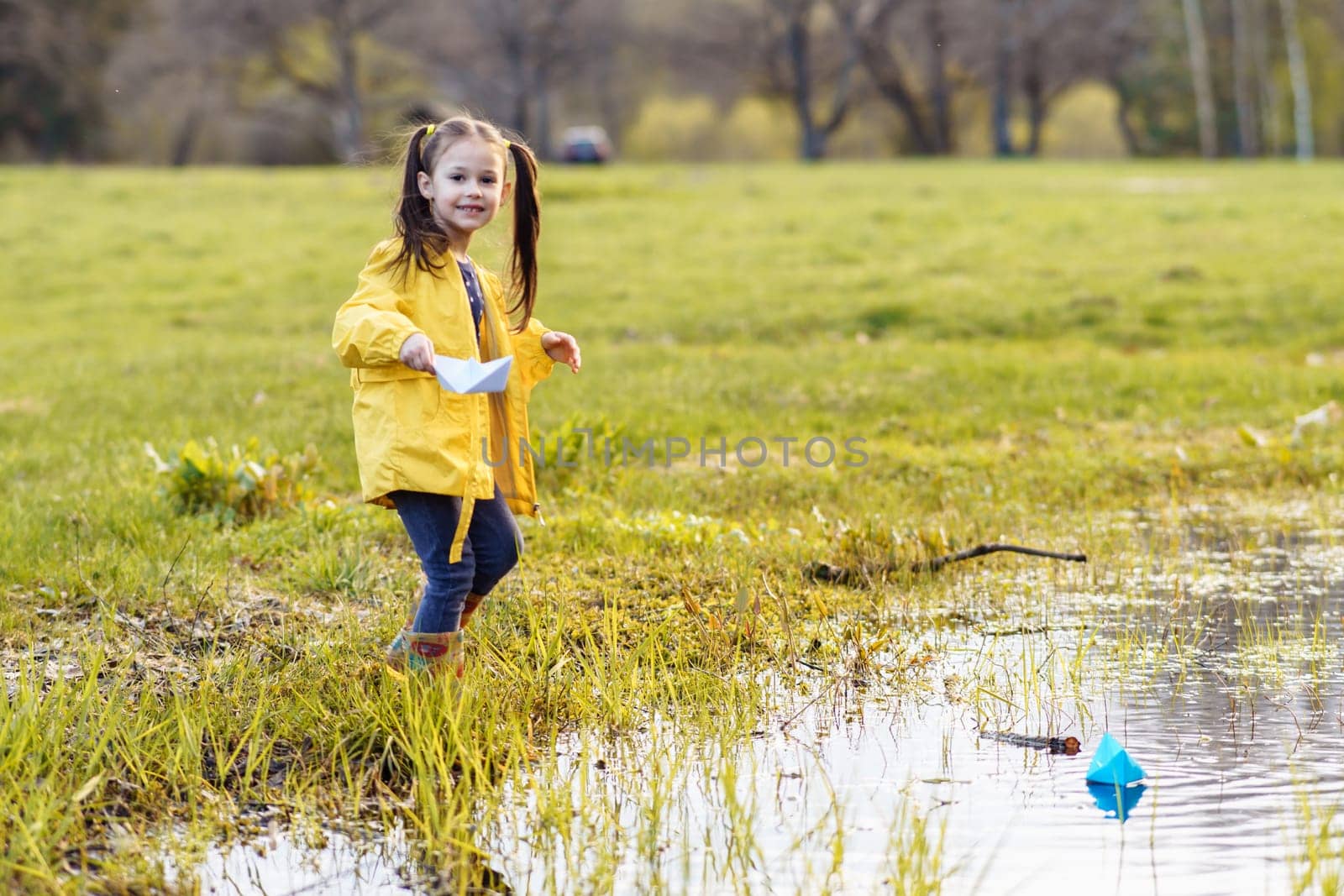 Portrait of gorgeous little girl, adorned in yellow jacket and rubber boots, holding white paper ship in hand and looking at camera with shiny smile, preparing to launch boat on puddle in autumn.