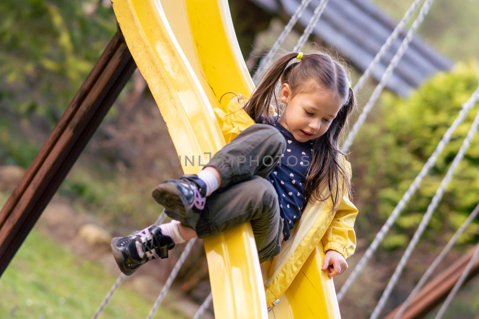 Active little girl with two ponytails sitting on bright yellow slide at playground, looking on way and carefully sliding down, spending funny leisure outdoors on warm autumn day.