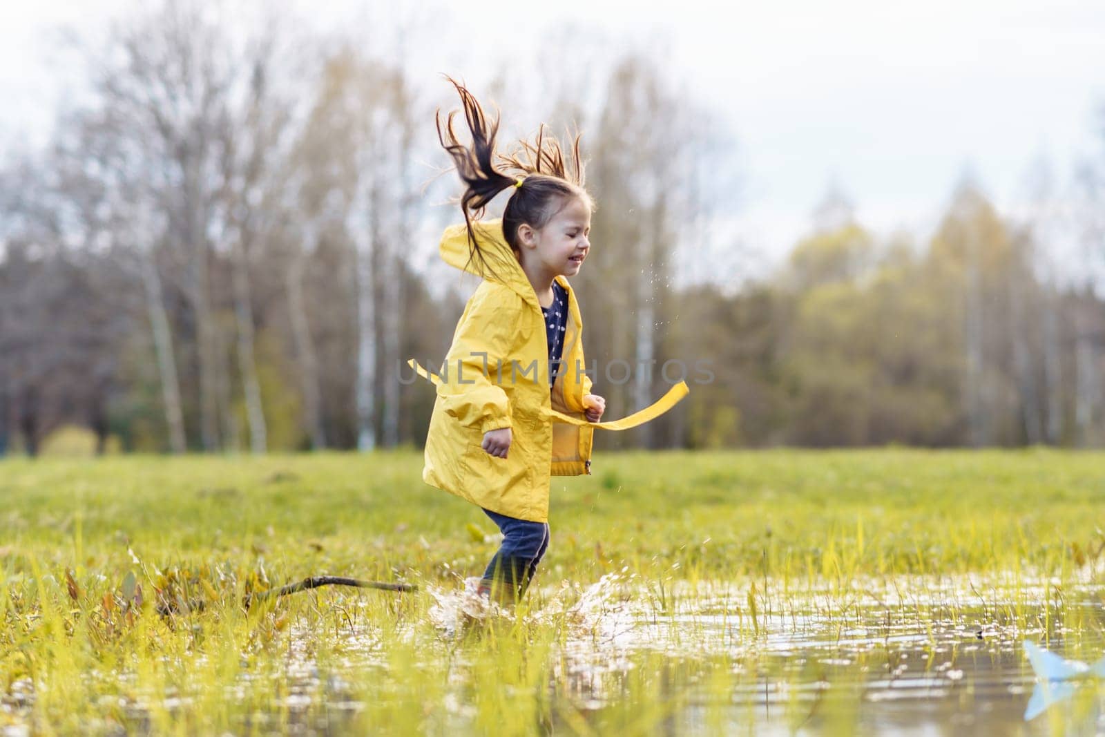 Adorable little girl, wearing yellow jacket, jumping on puddle, playing in pool in green lawn on autumn day. Female child with two ponytails, in rubber boots, closing eyes and splashing water.