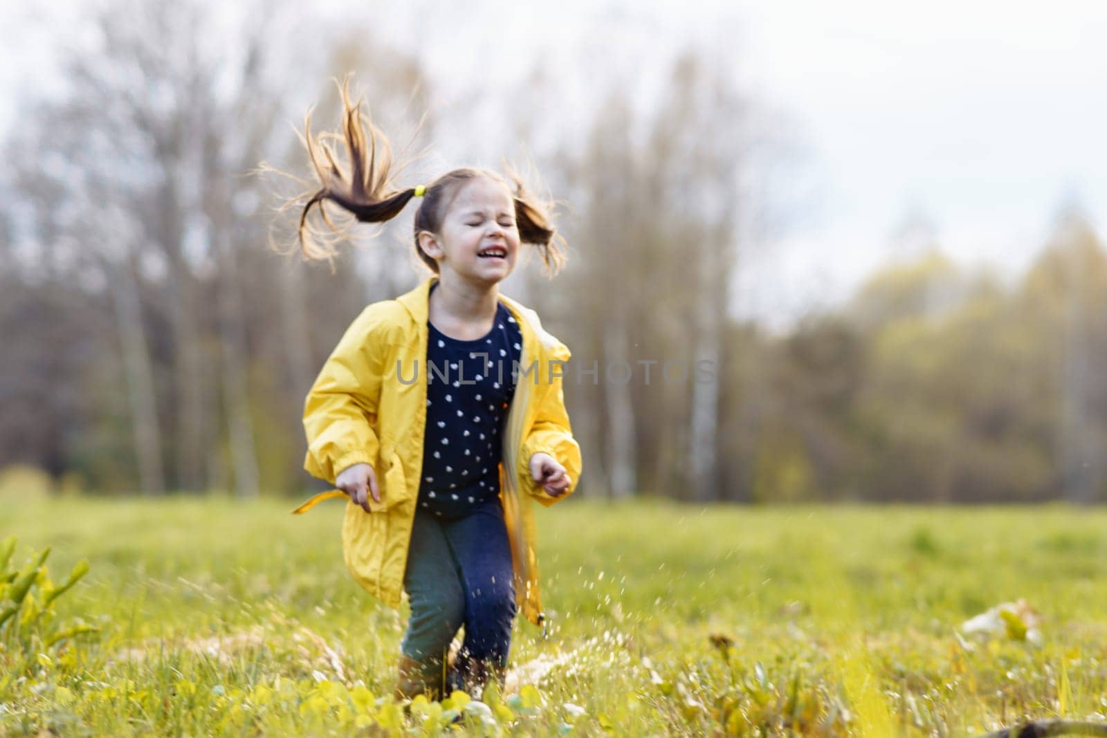 Gorgeous child in yellow jacket and rubber boots squinting eyes and jumping on puddle, playing with water on green site after rain. Little girl with ponytails splashing pool and enjoying leisure.