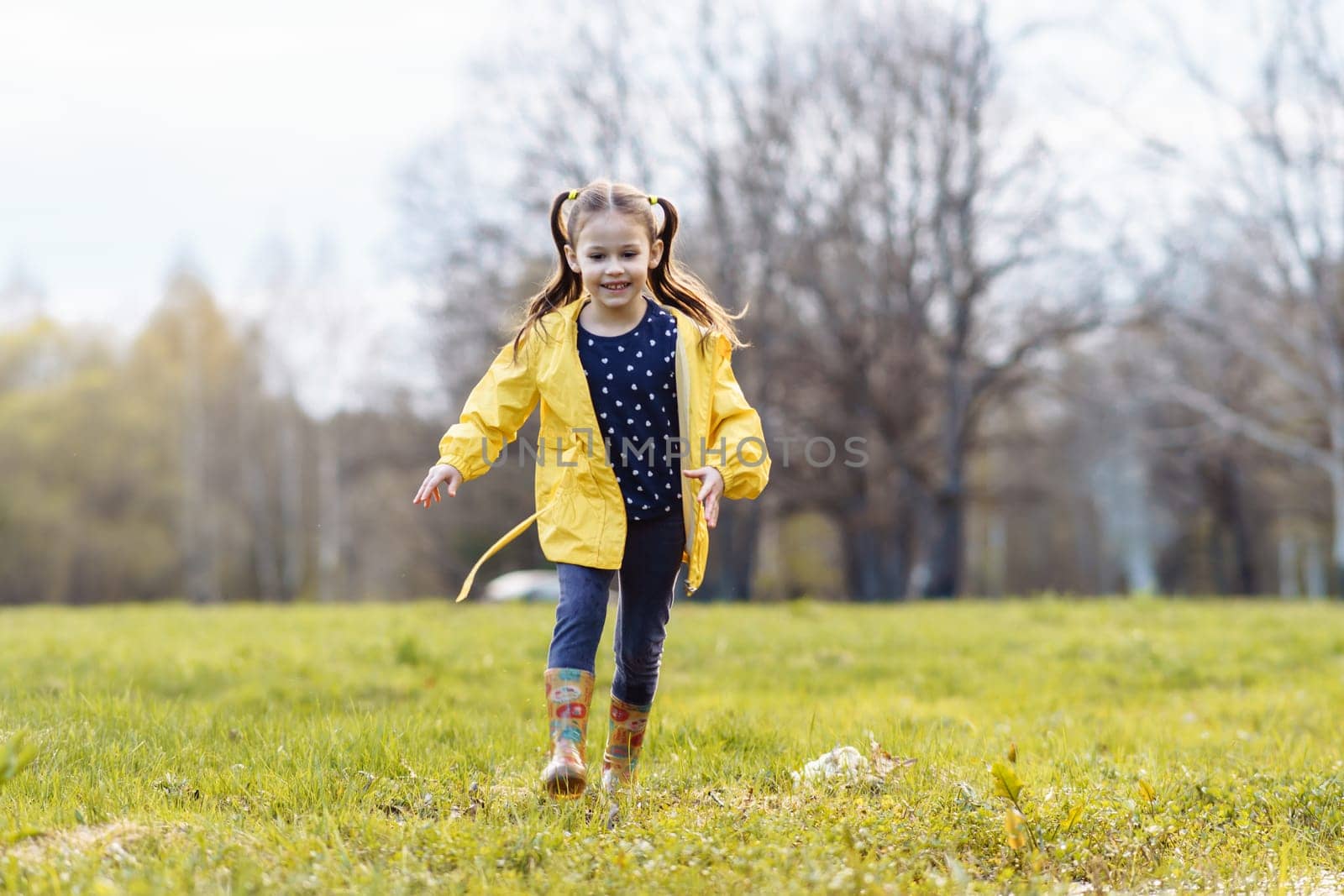 Loveable little girl with two ponytails, wearing yellow jacket and rubber boots, strolling across green site on autumn day, looking down with smile and eyes full of curiosity and carefree.