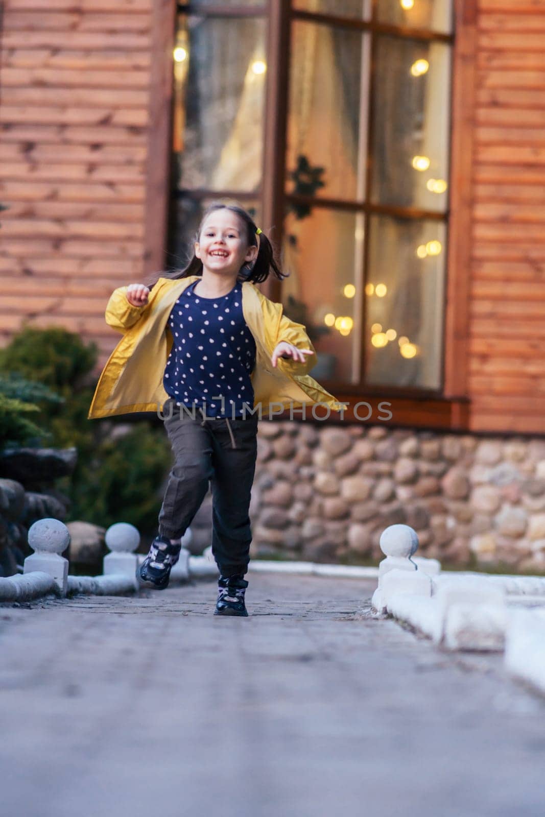 Glorious little girl with brown hair in two ponytails, in yellow jacket running from house and looking at camera with happy smile. Child going to walk on fall day, presenting funny leisure. Vertical.