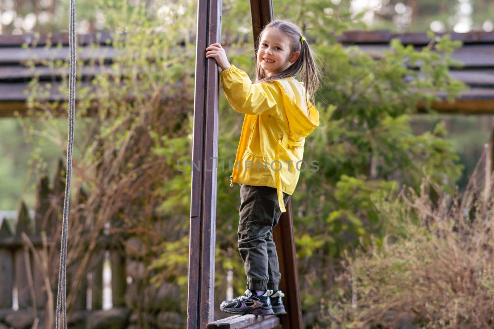 Glorious little girl, adorned in bright yellow jacket and black pants, standing on construction at playground, holding boards and turning head over shoulder, looking at camera with happy smile.