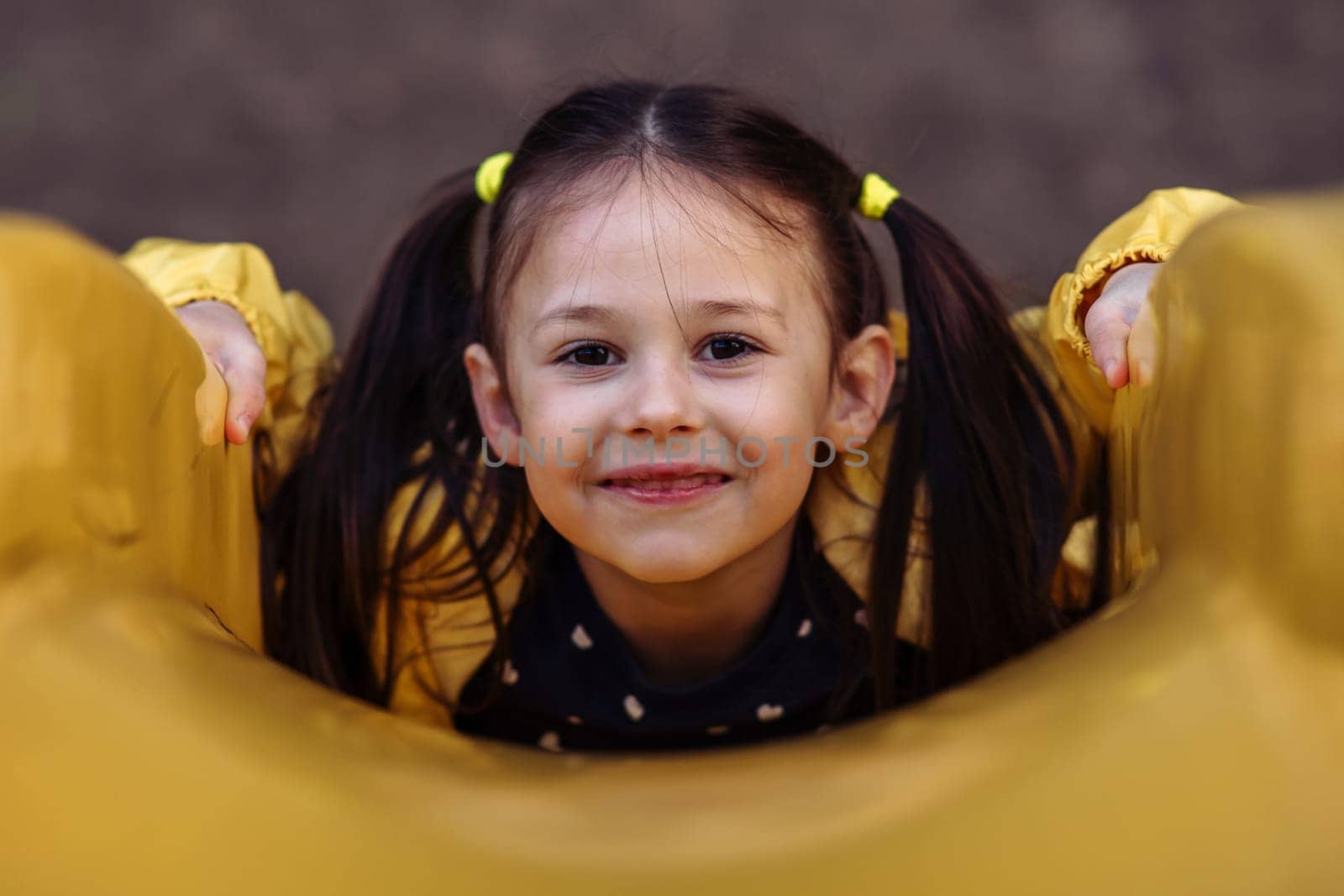 Portrait of charming little girl with brown hair tied up in two ponytails looking at camera and smiling while lying on stomach on yellow slide, having delightful walk at playground on autumn day.