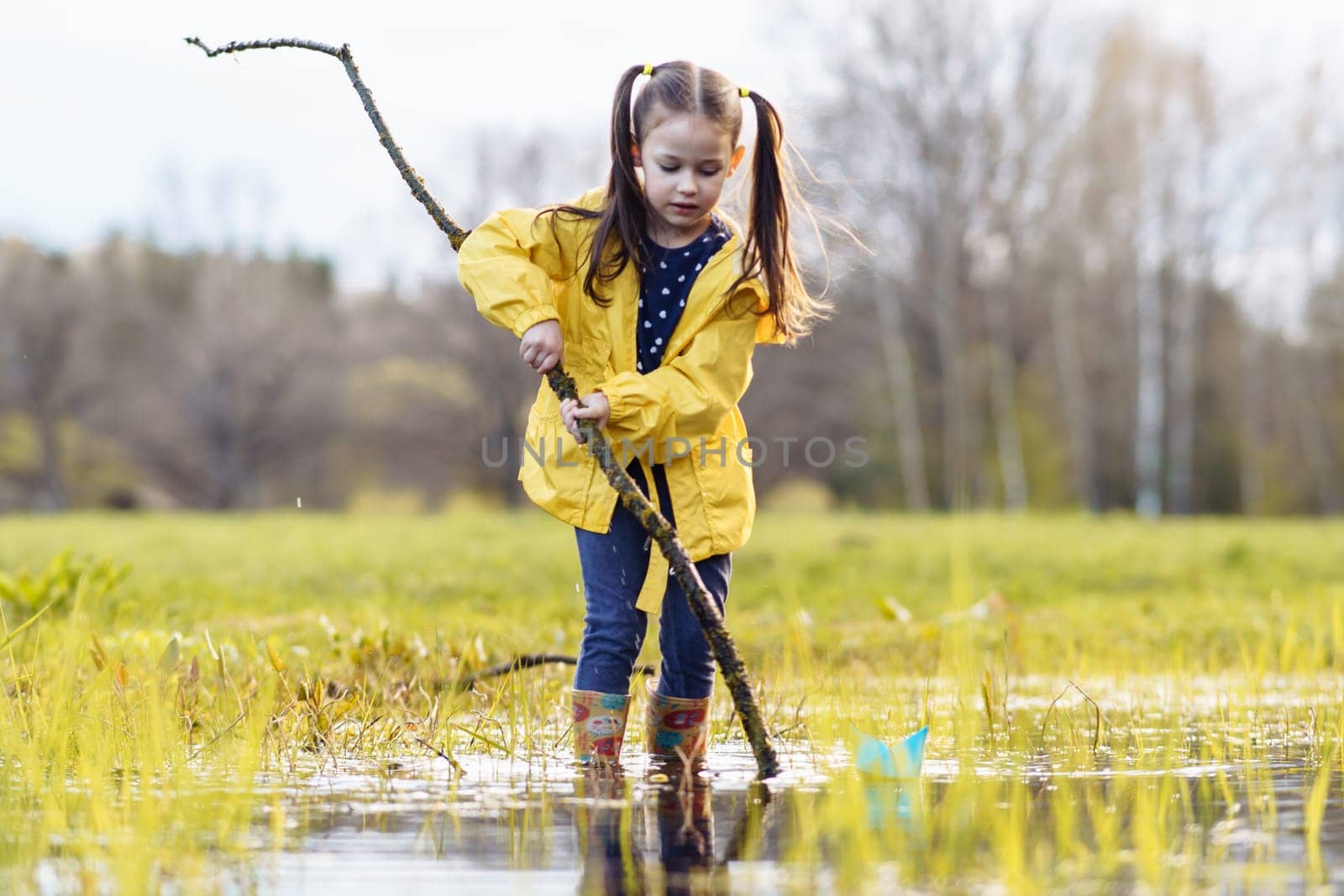 Concentrated little girl, wearing yellow jacket and rubber boots, holding long branch in hands while standing in puddle and looking down with focused eyes, preparing for playing with paper boats.