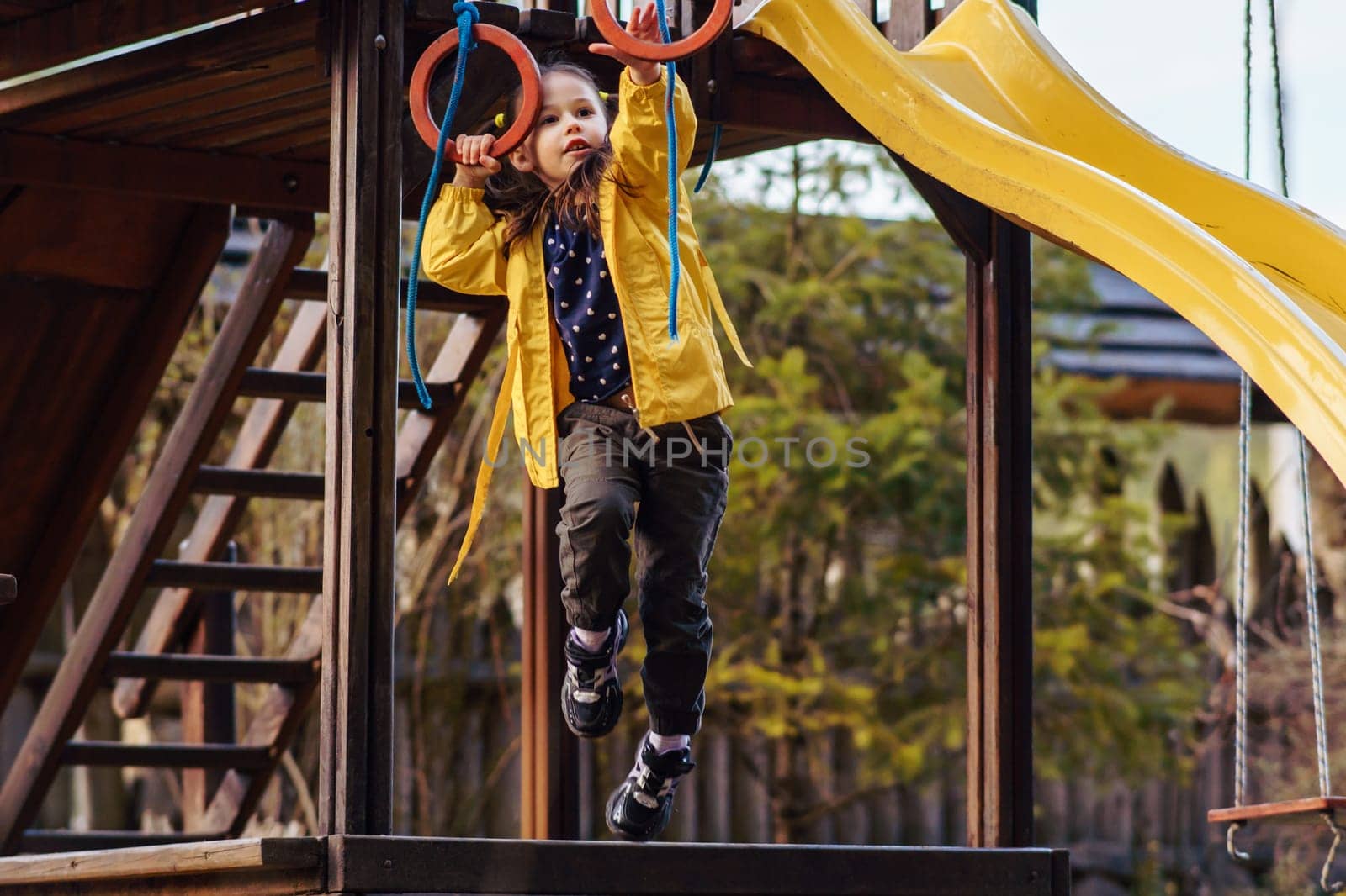 Energetic little lady, wearing bright yellow jacket, recreating at playground, climbing on red rings and enjoying leisure. Child holding equipment with hand and attempting to hang on small hoops.