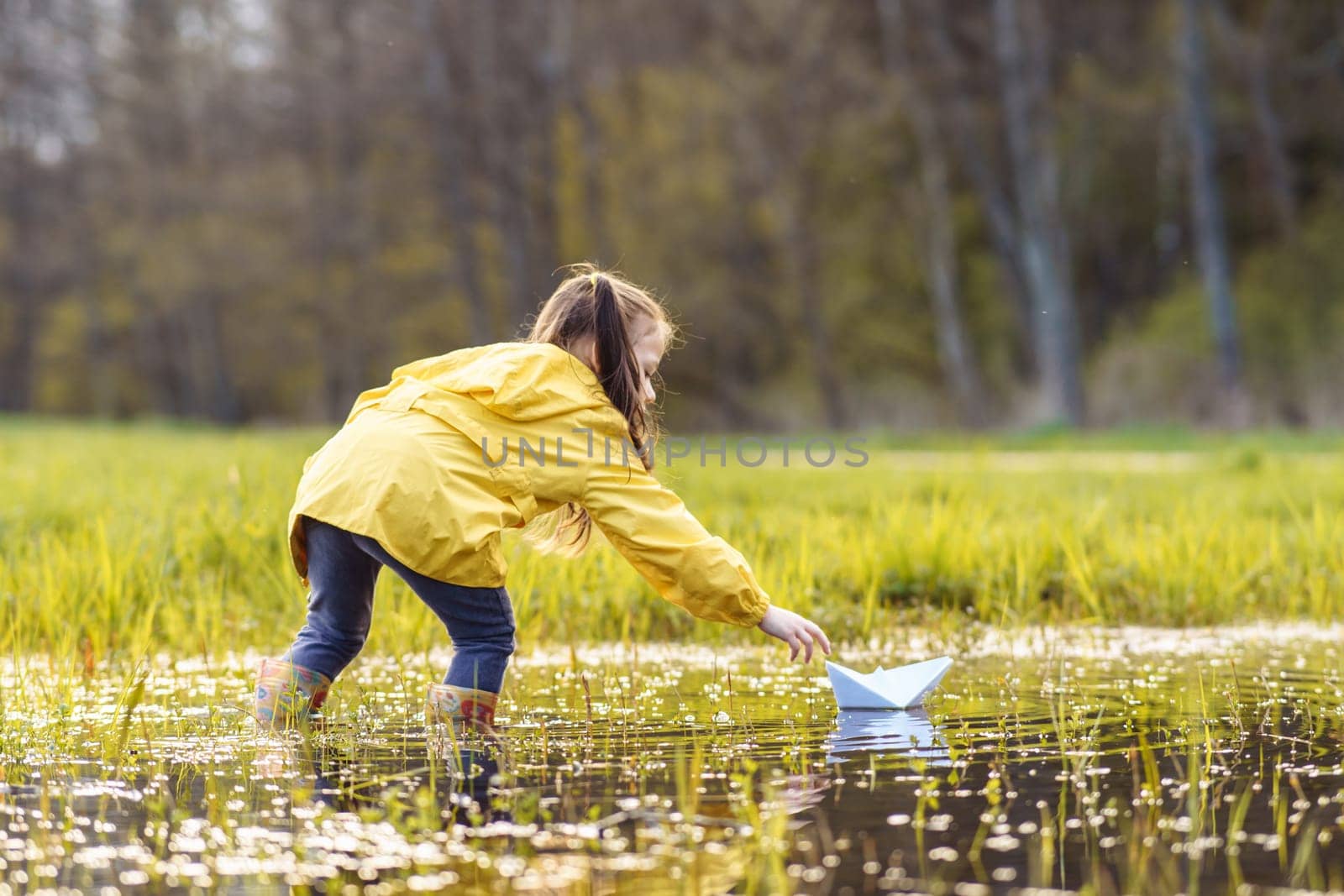 Rear view of little girl, wearing yellow jacket and rubber boots, standing in puddle and bending over closer to white paper ship, stretching arm trying to touch light boat, swimming on water.