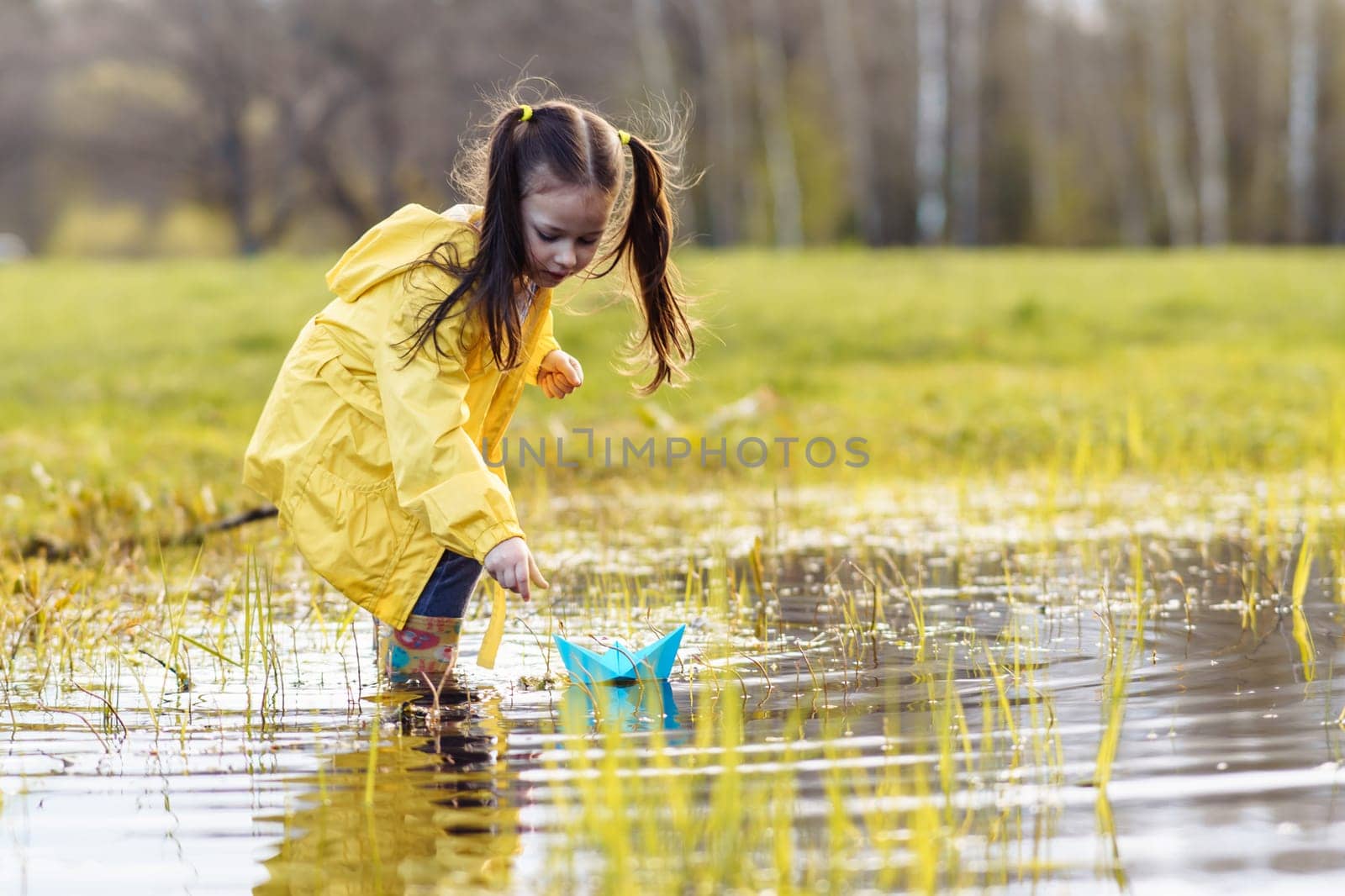 Delightful and calm child, wearing bright yellow jacket and rubber boots, standing in puddle in green lawn, bending over, stretching arm forward and launching blue paper ship, playing on autumn day.