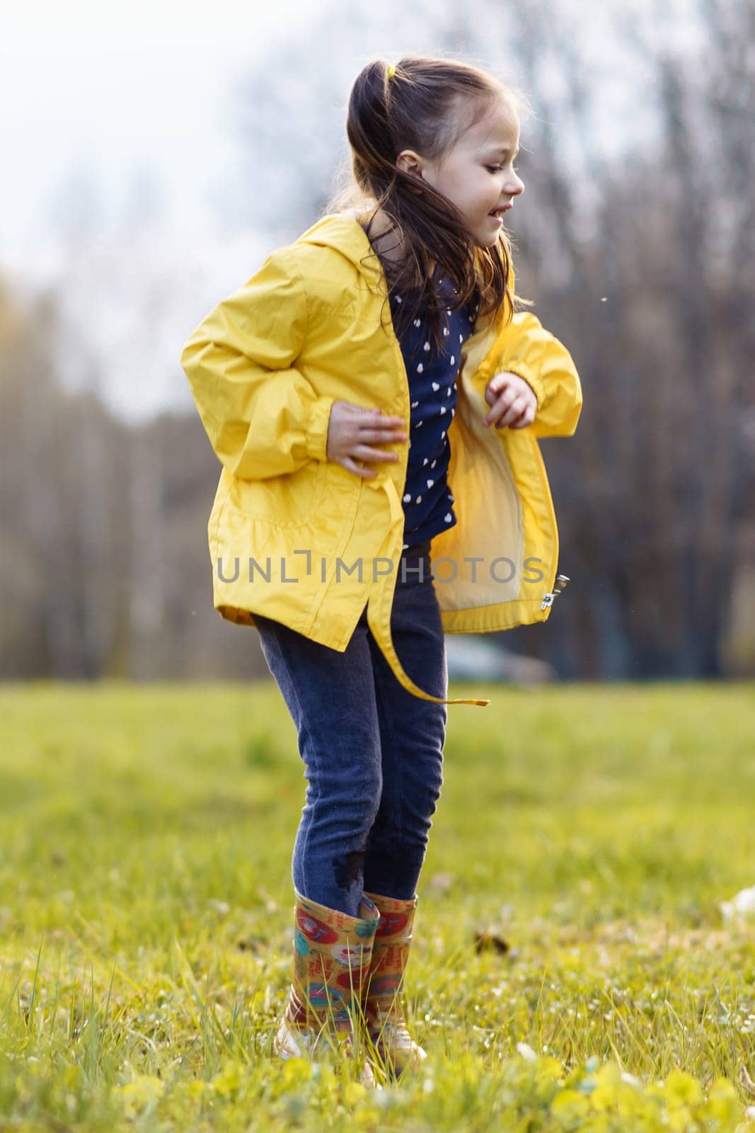 Joyful little girl, wearing yellow jacket and rubber boots, jumping up while standing in puddle on green lawn, looking down with carefree expression and enjoying autumn day after rain. Vertical.