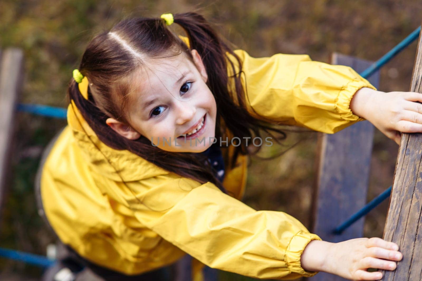 View from above of adorable little girl with two ponytails, adorned in bright yellow jacket, climbing on staircase constructed at playground and looking at camera with charming and shiny smile.